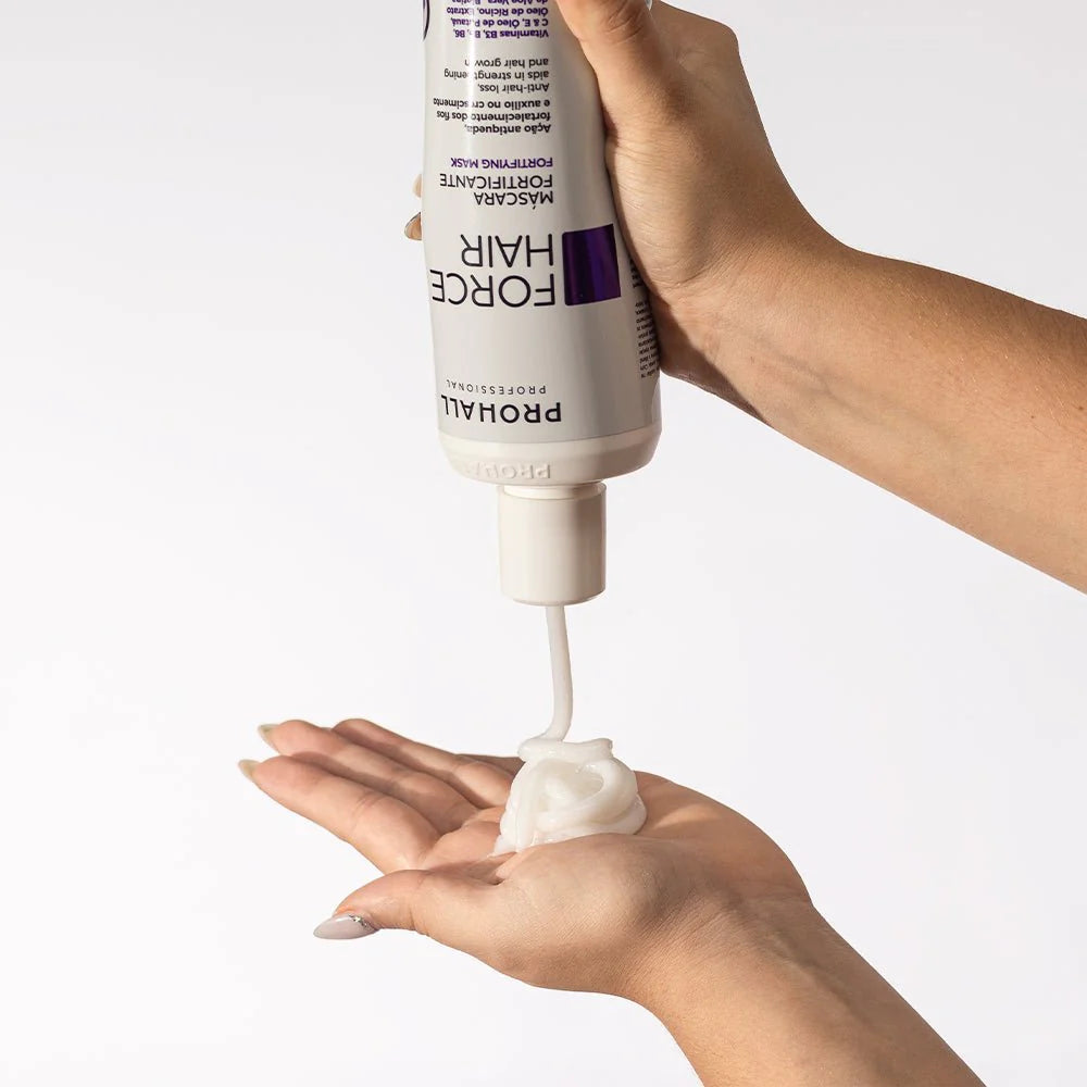 Hand holding a bottle of Prohall Force Hair product with cream being dispensed onto another hand against a white background.
