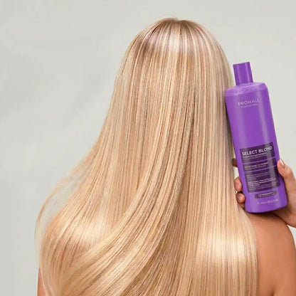 Person with long blonde hair holding a purple bottle of hair care product against a gray background