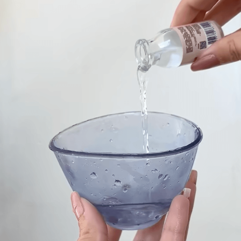 Person pouring liquid from a bottle into a glass bowl on a plain background